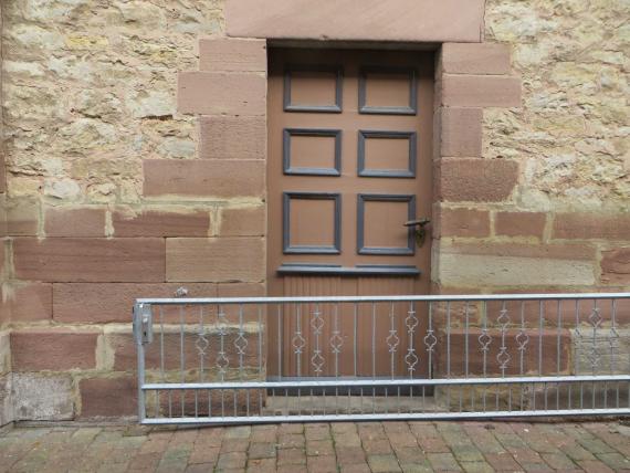 Frontal view of the narrow wooden door walled all around by red sandstone, where windows used to be. The door has six small black-framed wooden windows in the upper part and has a fresh coat of brown paint.