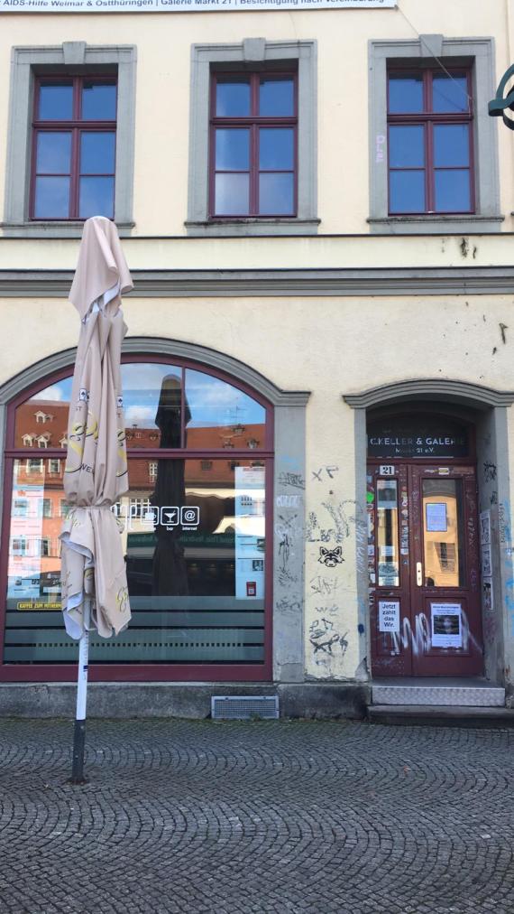 Auf dem Foto ist ein helles Gebäude zu erkennen. Links neben der Eingangstür sieht man ein großes Schaufenster. In der ersten Etage über Tür und Schaufenster lassen sich drei normale Fenster finden. Vor dem Haus steht ein zusammengebundener Sonnenschirm.