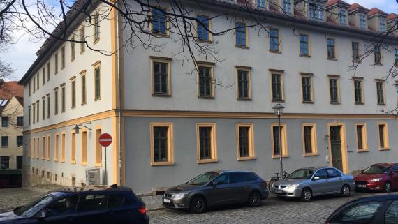 Foto von einem großem Wohnhaus. Der untere Teil des Hauses ist dabei dunkler als der obere Teil. Das Haus besitzt in jeder der drei Etagen sehr viele Fenster . Im Vordergrund stehen ein paar Autos am Straßenrand.