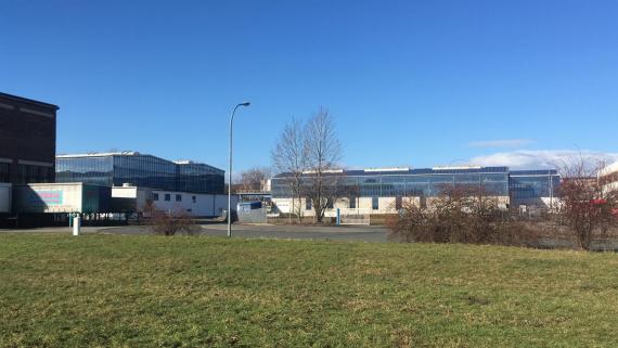 Das Bild zeigt einen großen Hallenkomplex mit zwei großen Fabrikhallen. Linkerhand. Es steht links eine Halle von schräger Ansicht, während zentral eine Seite der anderen Halle abgebildet ist. Außerdem beinhaltet das Foto eine große Himmel- und Wiesenfläche.