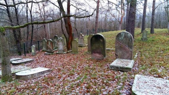 View of cluster of gravestones on trees at the lowered part of the cemetery, near the fence opposite the entrance. The gravestones are covered with a thin layer of snow.