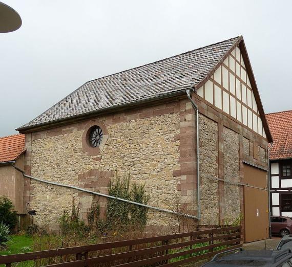 View of the synagogue from the main street. You can see a stone building with half-timbered elements, which has a large helter-skelter gate facing the street. On the left side, in the direction of a garden, a small round window is incorporated into the wall.