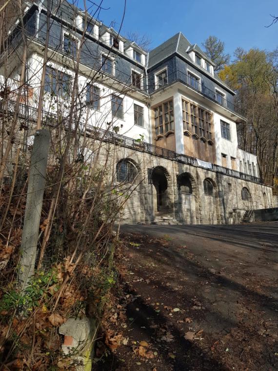 The photograph shows the Villa Simson located on the Suhl Cathedral Hill. Brushwood protrudes into the picture at the left edge. The first floor of the villa is made of stone, the entrance door is set into a round arch. The second floor is plastered white and has rectangular windows. A steel railing encloses the narrow terrace. Slate slabs line the second as well as the third floor, in which, unlike the other floors, there are no windows. They raise