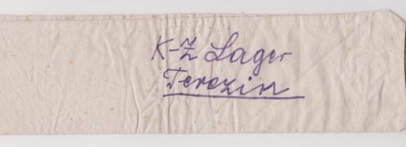 Man sieht eine Armbinde aus hellem Stoff mit dem handschriftlichen Schriftzug "K-Z Lager Terezin". Die Armbinde gehörte Clara Spaleck.