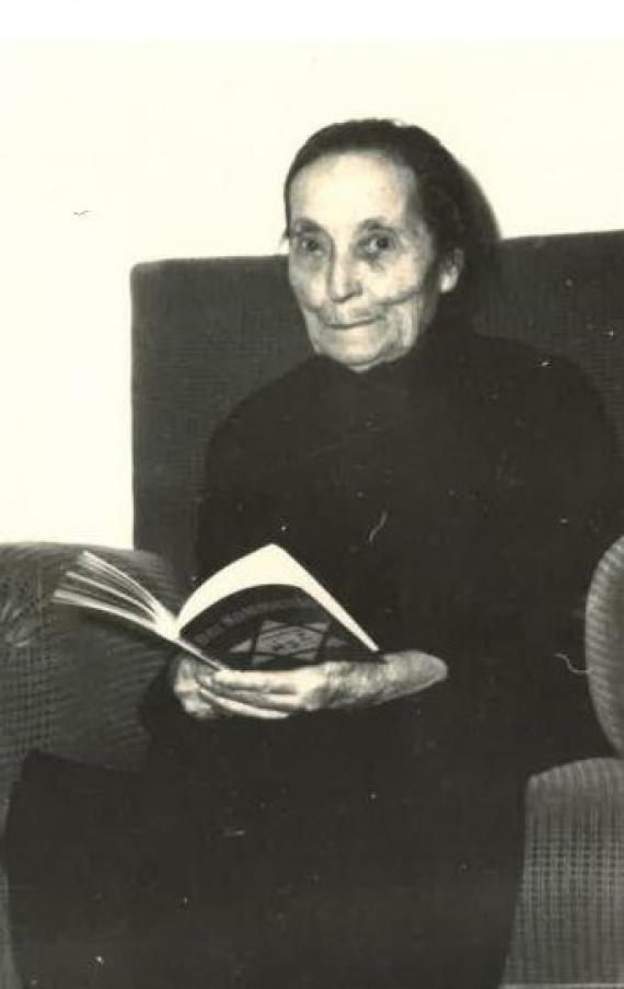 A black and white photo of Clara Spaleck. She is sitting on a sofa in a dark dress and has a book in her hands. The aged woman smiles slightly into the camera.