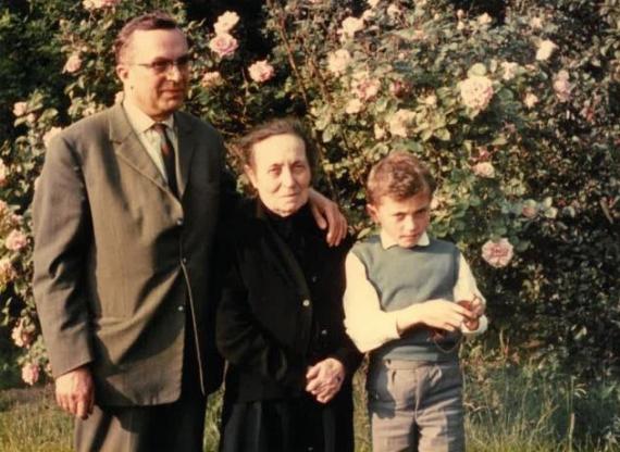 Zu sehen sind drei Personen, die vor einer blühenden Hecke stehen. In der Mitte steht Frau Clara Spaleck (in betagtem Alter, schwarzes Kleid), links steht ihr Sohn Siegfried Spaleck (hellbrauner Anzug) und rechts ein kleiner Junge (weißes Hemd mit grünem Pullunder).