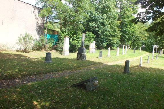 View of the cemetery. Some gravestones. In the background bushes and trees. And the back wall of a house left of center.