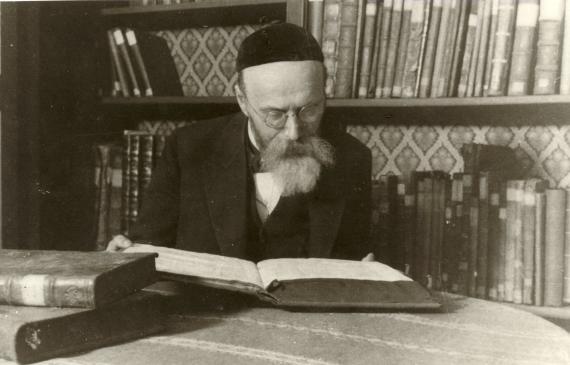 Rabbi Dr. Unna, seated at a table, bent over an open folio, two closed ones lying next to it. In the background a wall of books.