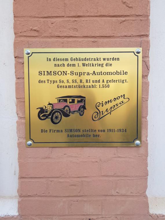 The photograph shows a commemorative plaque attached to a production building for the successful "Simson Supra" vehicle. The golden commemorative plaque reads: "SIMSON Supra automobiles of the types So, S, SS, R, RI and A were manufactured in this building wing after World War 1. Total number of units: 1,550. The SIMSON company manufactured automobiles from 1911 - 1934." Also shown is a drawing of the car.