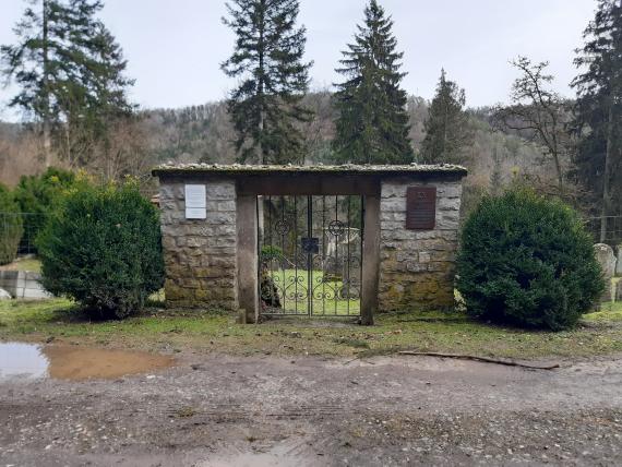 The entrance gate consists of two natural stone columns and has a simple roof. The two gate wings are made of iron. The rest of the cemetery is fenced with wire mesh.