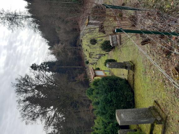 Blick über den Zaun des Friedhofs. Rechts erkennbar das Tor. Mittel und links die Grabsteine. Deutlich erkennbar ist die Informationstafel.