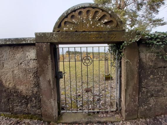 The entrance gate is located in the overman-high wall. The archway with Hebrew inscription towers above the wall. The gate wing made of tinned steel is decorated with a Jewish star. Through the grille one looks at the cemetery.