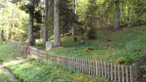 Der Firedhof liegt mitten im Wald unter alten Bäumen. Er ist von einem kniehohem Lattenzaun umgeben.