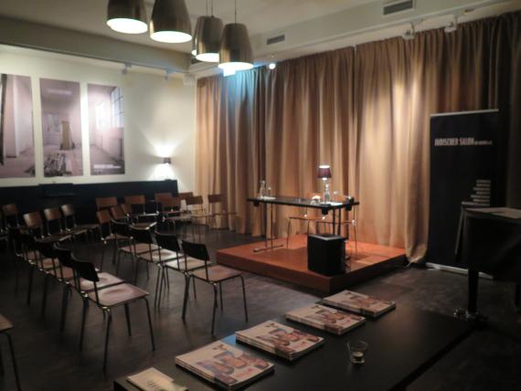 A table with two chairs behind it. On the table water carafes, glasses, a small reading lamp with a red shade. The table stands on a small podium. Lined by beige curtains behind the table. In front of it several rows of chairs.