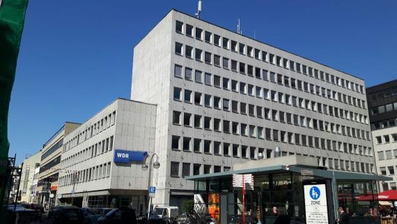 Großes, graues, kastenförmiges, mehrgeschossiges Neubau-Gebäude mit WDR-Logo vor einem blauen Himmel. 