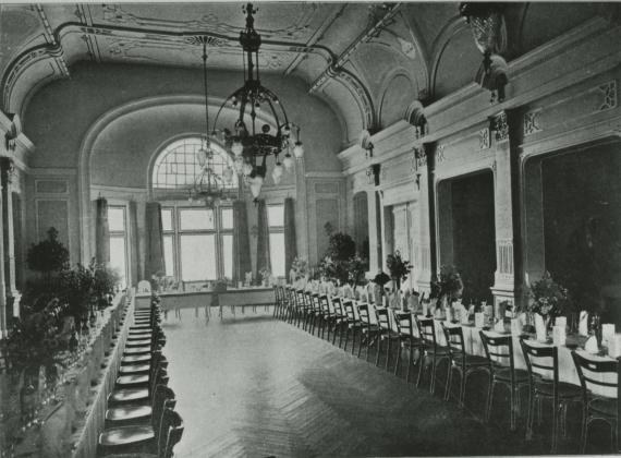 Ballroom on the second floor of the lodge house at Hartungstraße 9-11. A long continuous row of tables on all sides of the room, plus simple wooden chairs in front of each seat. White tablecloth. At each place a table setting, plus several vases with flowers. A chandelier hangs in the center of the room. A second one hangs above the row of tables at the head end. At the back of the room a large window.