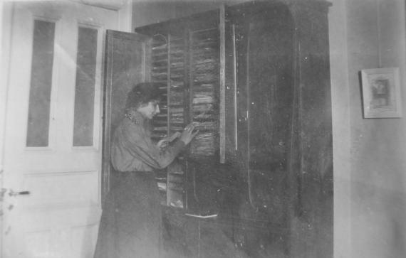 Ida Dehmel stands in front of an open, almost door-high cabinet. In it are various compartments, each containing many sheets of paper: The Dehmel archive. To the left of Ida Dehmel is a white door and to the right of the cabinet hangs a small picture on the wall.