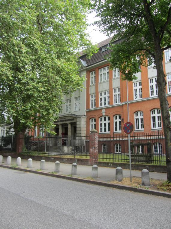 Außenansicht der ehemaligen Talmud-Tora-Schule von der gegenüberliegenden Straße aus, flankiert von zwei Bäumen. Das Gebäude ist von einem Metallzaun umgeben. Zum Eingang führt eine kleine Treppe hoch. Der Eingang selbst ist antiken Säulen (insgesamt drei Stück) nachempfunden. Darüber steht in großen, schwarzen Buchstaben: Talmud-Tora-Realschule. Darüber in kleineren Buchstaben: 1911 - 1939.