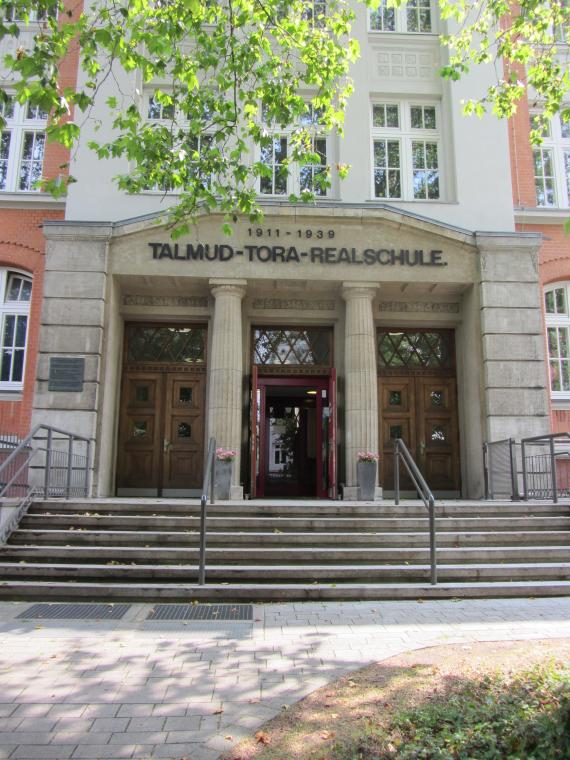 Entrance portal Carlebach school, six steps lead up. The portal has two columns in the center, which are modeled on ancient columns. In total there are three large entrance doors made of dark wood. The inscription above the portal reads: "1911 - 1939 | Talmud Torah Secondary School".