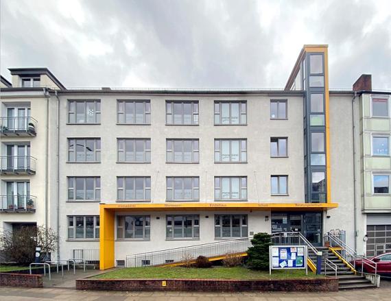 Schämferkampsallee, today a Montessori school. The building has a window front of four by four windows. In total, it has a first floor and three other floors. A yellow roof is installed from the second window from the left on the first floor. On it is written: "monadrei, Montessorie, children's house, school, counseling center". The roof merges into a projecting bay window that goes over the three upper floors and merges into a fourth attic.