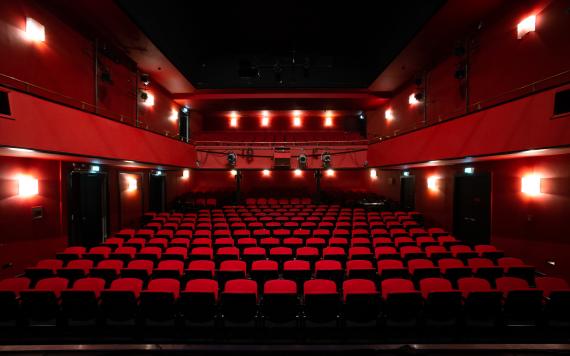 View into today's theater hall of the Hamburg Kammerspiele. View from the stage to the folded-up rows of seats. All covered with red velvet. In general, the hall is red. In the background you can see the upper floor of the theater. Lamps on the sides and back of the wall discreetly illuminate the room.
