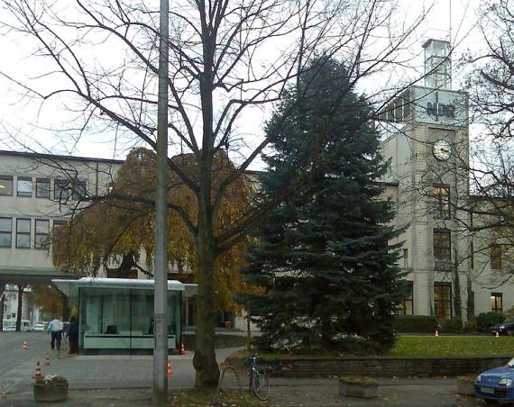 Headquarters of the North German Broadcasting Corporation. An elongated building complex with a prominent four-story tower and glass roof. On it in dark blue, large letters NDR. Below it an analog clock (without numbers). In front of it a fir tree, a weeping willow and in front of it another tree without leaves. On the left is a glazed gatehouse.