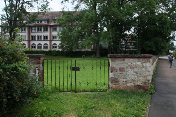 Eine halbhohe Mauer. Ein eisernes Tor. Dahinter auf einer Wiese Grabsteine.