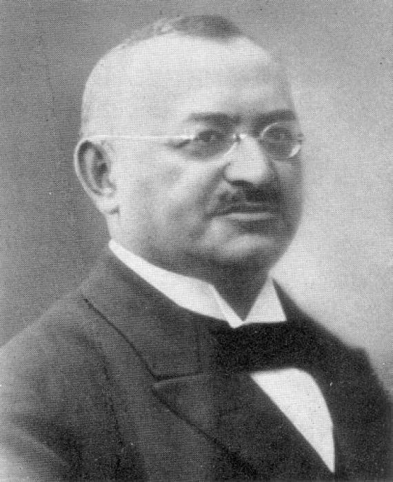 Black and white portrait of Max Beermann in half profile. Max Beermann is a middle-aged man with glasses, a mustache and fair skin. He wears a dark jacket, a white shirt and a black bandage over his collar.