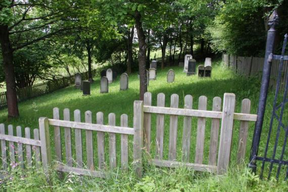 A wooden fence with vertical slats. Gravestones under trees. On the right side of the fence a gate.