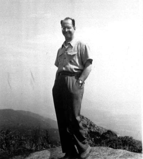 Black and white photograph of Hans Schiff in relaxed pose on a hill (full body photography) with mountains in background