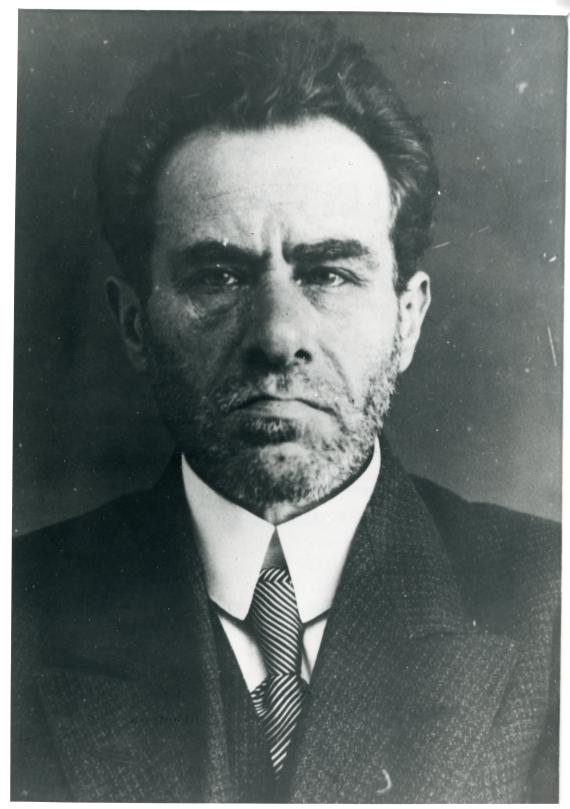 Frontal shot in black and white. A man with serious look. His dark hair is combed back. He looks straight ahead.