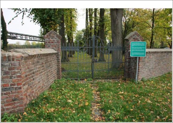 A green iron gate. A wall on both sides. Through the gate you can see a meadow, trees, gravestones.