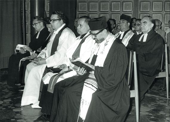 Ein schwarz-weiß Bild. Mehrere Männer in religiösen Gewändern unterschiedlicher Konfessionen sitzen gemeinsam in Stuhlreihen.