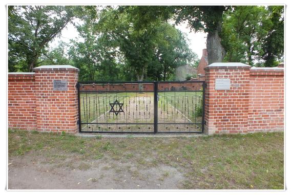 Ein hohes und breites Tor aus Eisenstangen. Seitlich gehalten von zwei gemauerten Pfeilern. Dahinter der Friedhof.