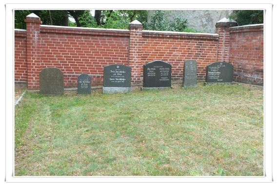 Several dark stones in a row in front of the cemetery wall. High wall of red bricks.