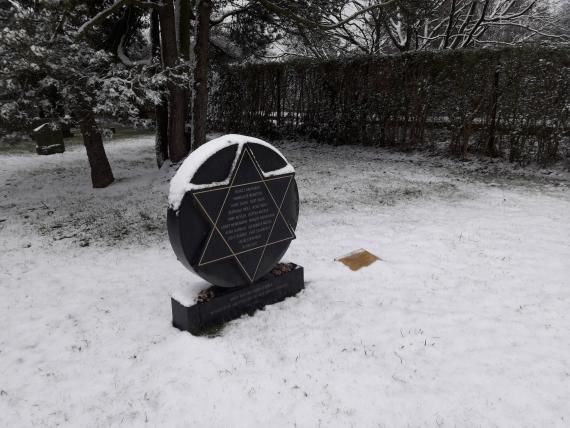 The memorial stone made of dark, smooth stone consists of a round flat plaque, with a Star of David in it. The names of the deported Jewish children from Grevenbroich are carved on the front of the stone. The memorial stone stands on a rectangular pedestal. To the right of it is a small plaque with information about its installation on the ground. The stone stands on snow-covered grass. In the background two trees and the hedge surrounding the cemetery can be seen.