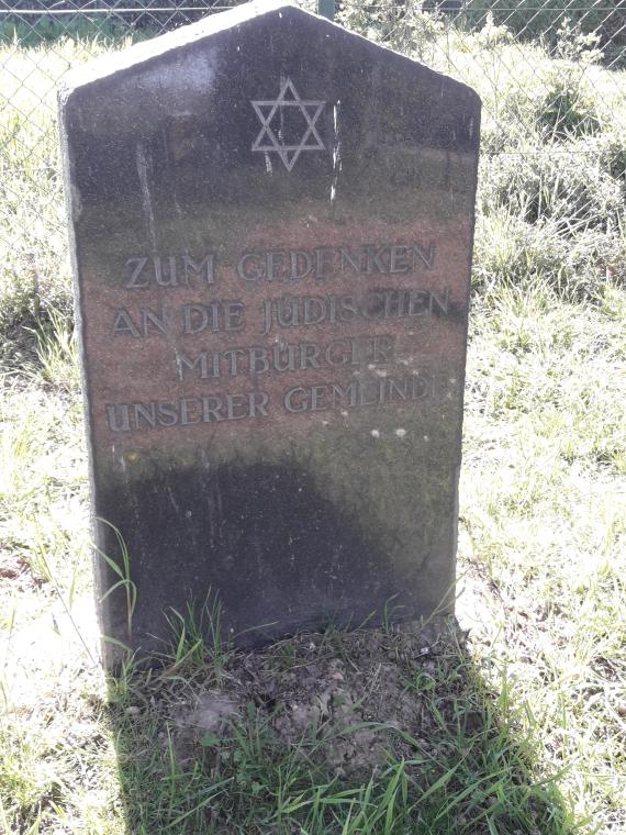 The memorial stone is made of dark, smooth stone and has approximately the size of a simple gravestone. It is rectangular and tapers to a triangle at the top. In its pediment is engraved a Star of David, below it the inscription "In memory of the Jewish fellow citizens of our community". The memorial stone stands in a row next to the other gravestones on the right side of the central path. Behind it, the low wire mesh fence surrounding the cemetery can be seen.