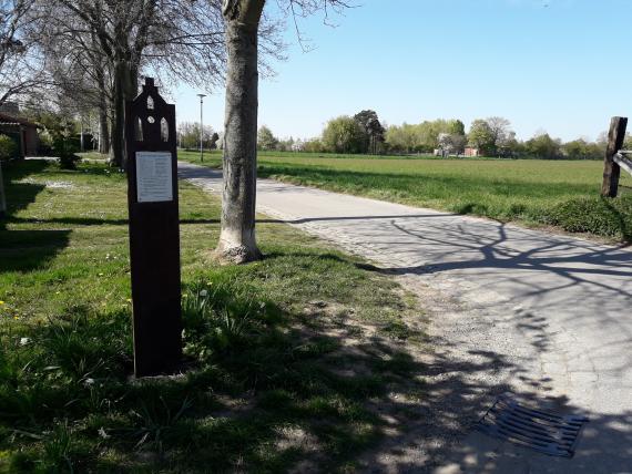 In der Mitte des Bildes verläuft die Straße Am Fleckenweiher. Links der Straße steht die Gedenkstele, die an den alten jüdischen Friedhof erinnert. Dahinter stehen entlang der Straße im regelmäßigen Abstand mehrere Bäume. Rechts der Straße liegt das Grundstück, auf dem sich der alte jüdische Friedhof befand. Heute stehe dort keine Grabsteine mehr. Die Wiesen rechts des Weges sind weitläufig. Im Hintergrund sind mehrere Bäume zu erkennen. 