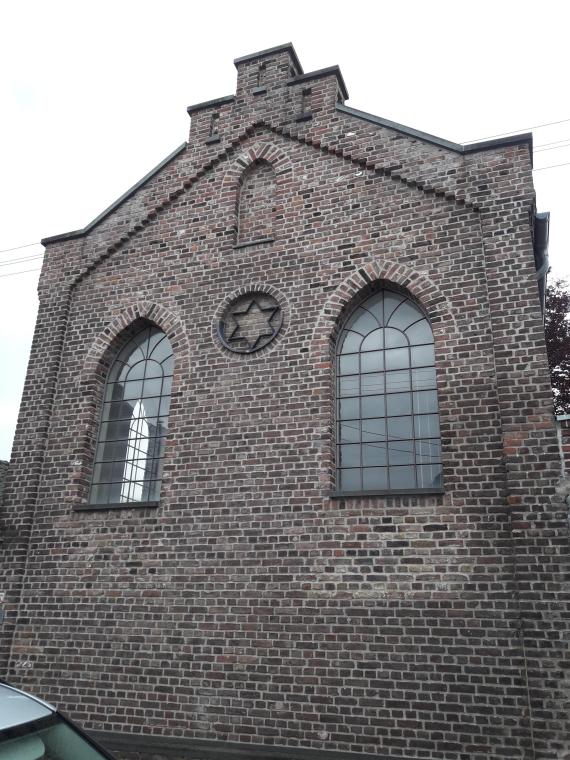 Gesamtansicht der Fassade. Durch die beiden großen Fenster und die fehlende Zwischendecke erscheint der Betraum im Inneren recht weit und hell, obwohl er nicht groß ist. Auf seiner Rückseite befindet sich auf der Höhe des ersten Stocks eine kleine Frauenempore mit gesondertem Zugang. 