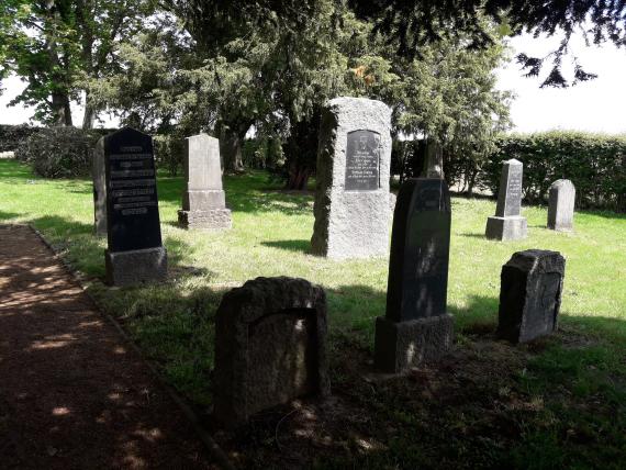 The photo shows the gravestones in the middle part of the cemetery on the right side of the path. On the grass the gravestones stand in several rows one behind the other. In their shape and size they are quite different. Some have been newly edged or otherwise repaired. In the background there is a big tree.