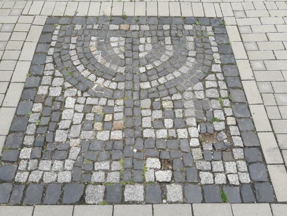 Auf dem Foto ist ein quadratisches Mosaik zu sehen, das in den hellgrau gepflasterten Gehweg eingelassen ist. Das Mosaik besteht aus mittelgrauen und dunkelgrauen kleinen Kopfsteinen. Die mittelgrauen Steine bilden den Hintergrund, während aus den dunkelgrauen Steinen eine Menorah sowie diese umgebende Ornamente gelegt wurde. Ein Teil der dekorativen Muster in der rechten unteren Ecke des Mosaiks ist beschädigt. An dieser Stelle fehlen Pflastersteine. 