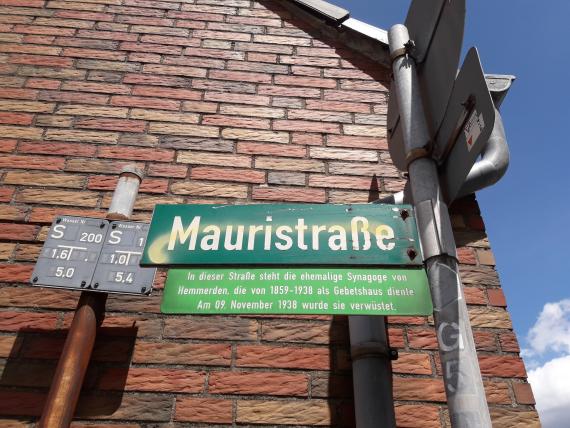 Unterhalb eines dunkelgrünen Straßenschildes mit der Aufschrift Mauristraße ist eine kleinere hellgrüne Informationstafel angebracht. Diese umreißt knapp die Geschichte der Hemmerdener Synagoge. 