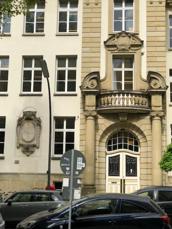 This photo shows the outside of a building with focus on a door with a balcony above. There is a sign saying knaben schule or boys' school to the left of the door.