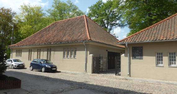 Two one-story houses with red tile roofs, in front a street with cobblestones and parked cars, behind them can be seen treetops. Between the houses is an entrance gate made of black metal.