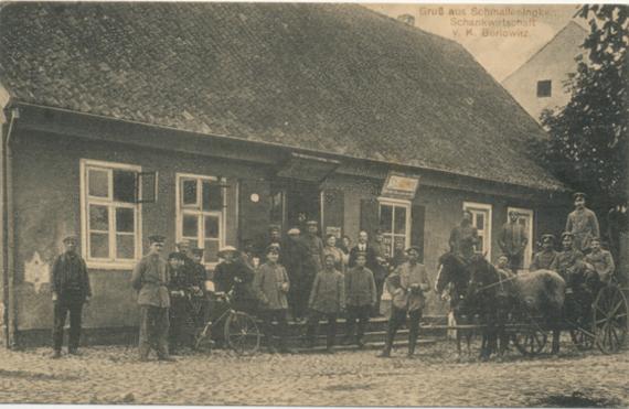 In front of one-story house there are many people, a cyclist and a carriage with two horses. Most of the people are wearing military uniforms and are male. Another uniformed man, possibly a letter carrier, holds the bicycle, in front of the open entrance in the middle are two women and a man with a tie.