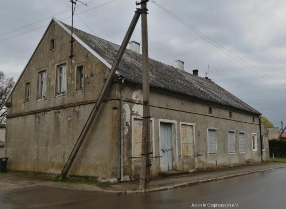 Plastered gray single-story gabled house on the corner of a street intersection. At the corner, in front of the house is a wooden pole with side support for power lines. Long side with six windows and a door, gable end has no windows below. Everything is locked and bolted. Street is asphalted - on the left in the side street you can see the beginning of cobblestones.