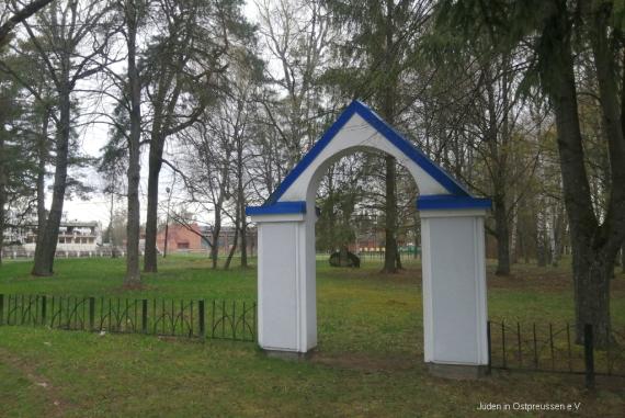 A white open archway with a blue roof marks the entrance to the area marked with a low black metal fence under trees. Through the gate one can remotely see the memorial stone for the cemetery. In the background one can see the neighboring sports facility.