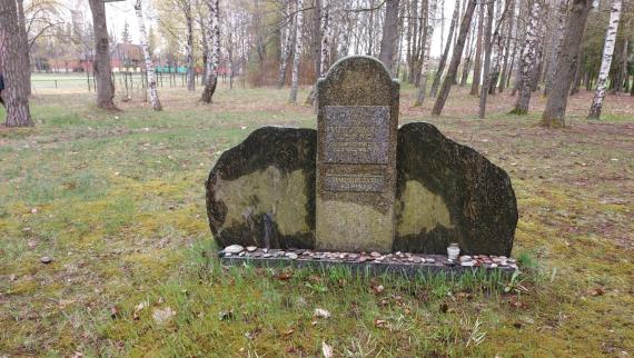 A flat pedestal, covered with loose pebbles. An upright tablet in the shape of a Jewish tombstone, rounded at the top. With two panels with inscriptions. Behind it a large erratic boulder of gray-green granite, which gives the whole an irregular round shape. Behind it trees of the adjacent forest and in the background parts of the sports field.  