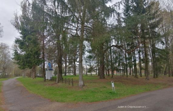 Exterior view of the cemetery seen from the corner of the asphalt road and the dirt road. On the left side there is the archway of the memorial place with the low fence, on the right side in the middle the area with very large and different coniferous trees. In the background modern buildings of the place shimmer through.