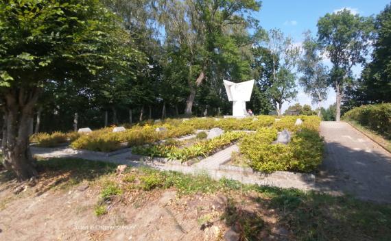 Außen sind hohe Bäume, links hinten eine Umzäunung. Man sieht ein rechteckiges Feld mit rechteckigen Betonunterteilungen, beflanzt mit niedrigen Büschen und es ragen stumpfe Betonelemente aus dem Boden. Ein mittelhohes weißes Denkmal steht leicht versetzt im Gräberfeld.